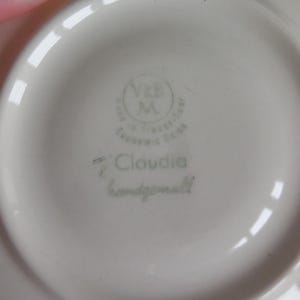 May include: Close-up of a white ceramic saucer with a light green stamp. The stamp includes the text "V&B M" inside a circle, with additional text around it. The word "Claudia" and "handgemalt" are also visible.