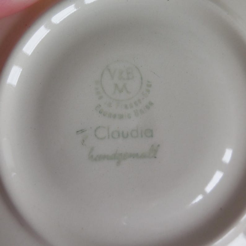 May include: Close-up of a white ceramic saucer with a light green stamp. The stamp includes the text "V&B M" inside a circle, with additional text around it. The word "Claudia" and "handgemalt" are also visible.