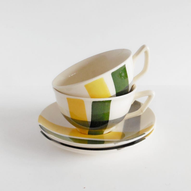 Peut inclure: Deux tasses à thé et soucoupes en céramique. Les tasses et les soucoupes sont blanches avec un motif de rectangles jaunes, verts et gris. Les tasses ont une anse. Les soucoupes sont empilées.