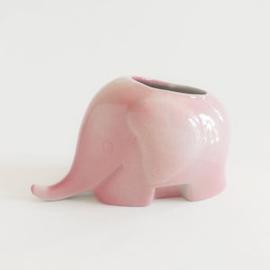 Puede incluir: Una maceta de cerámica brillante con forma de elefante en un suave degradado de color rosa claro. La figura del elefante presenta un cuerpo liso y redondeado con una trompa extendida hacia adelante y una amplia abertura en la parte superior.
