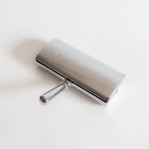 Op de afbeelding: Een gepolijst verchroomd metalen object met een rechthoekig, licht gebogen bovendeel dat is bevestigd aan een enkele, gladde, cilindrische handgreep die vanuit het midden uitsteekt. Het oppervlak is reflecterend en metaalachtig met een egale afwerking.