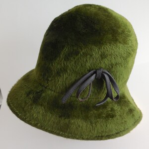 May include: A green, fuzzy cloche hat with a dark brown leather bow. The hat has a rounded crown and a slightly flared brim. The fur-like texture gives the hat a vintage look.