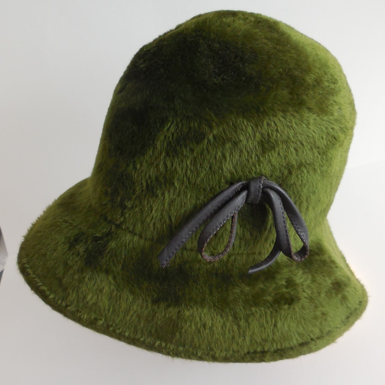 May include: A green, fuzzy cloche hat with a dark brown leather bow. The hat has a rounded crown and a slightly flared brim. The fur-like texture gives the hat a vintage look.