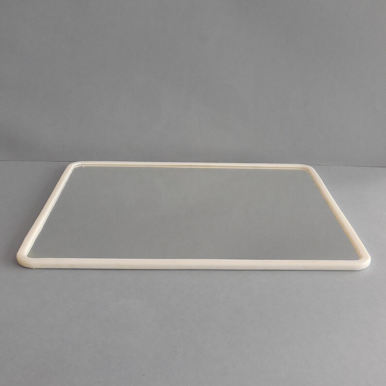 May include: A rectangular mirror with a rounded, off-white frame. The mirror's reflective surface is smooth and clear, set against a neutral gray background. The frame's corners are gently curved, creating a soft, modern aesthetic. The mirror is a decorative item.