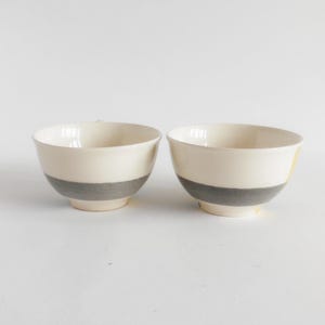 May include: Two ceramic bowls with a cream-colored upper section and a gray band around the middle. The bowls have a simple, modern design and are set against a white background. The bowls are likely for serving food.