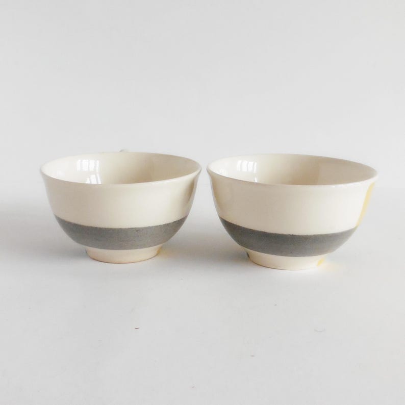 May include: Two ceramic bowls with a cream-colored upper section and a gray band around the middle. The bowls have a simple, modern design and are set against a white background. The bowls are likely for serving food.