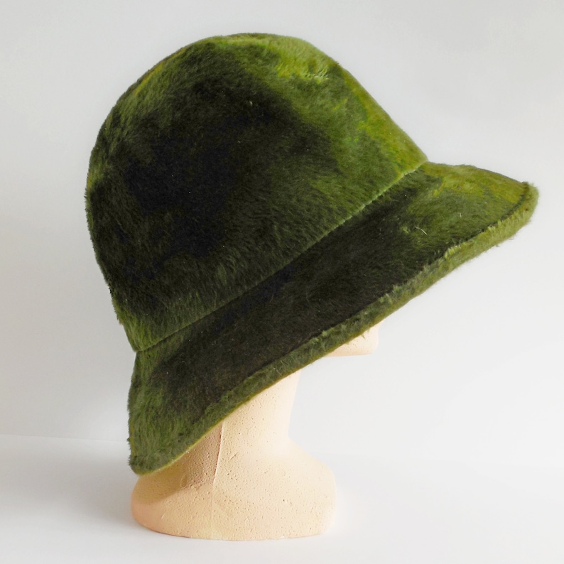 May include: A vintage green bucket hat with a soft, fuzzy texture. The hat has a rounded crown and a small, downward-sloping brim. The hat is displayed on a mannequin head against a white background. The hat's color is a deep, mossy green.