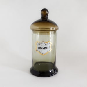 Op de afbeelding: Een vintage apothekerspot met een donkerbruine deksel en basis, en een heldere glazen body. De pot heeft een goud en wit label met het woord "Pyramidon". De pot wordt waarschijnlijk gebruikt voor het bewaren van medicijnen of andere items.