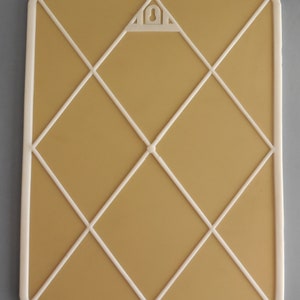 May include: Rectangular memo board with a cream-colored frame and a tan interior. White lines form a diamond pattern across the surface. A triangular hanger is at the top center. The board is designed for displaying notes, photos, or other items.