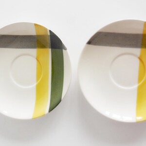 May include: Two white ceramic saucers with a central recessed circle. Each saucer features a design of vertical and horizontal stripes in yellow, green, and gray. The saucers are set against a white background.
