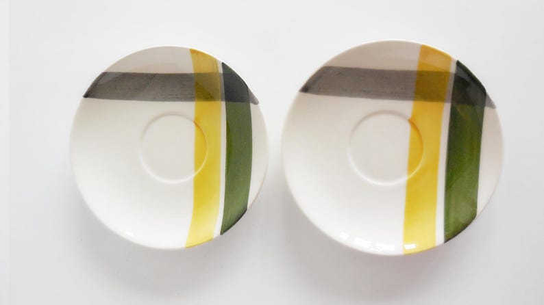 May include: Two white ceramic saucers with a central recessed circle. Each saucer features a design of vertical and horizontal stripes in yellow, green, and gray. The saucers are set against a white background.