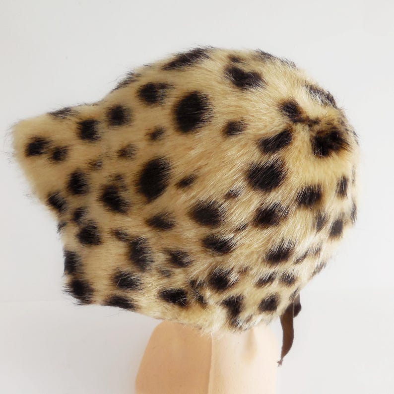 May include: A leopard print faux fur hat. The hat has a tan base with dark brown spots. It features a brim and a brown strap. The hat is made of a soft, plush material. A stylish accessory.