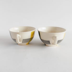 May include: Two white ceramic teacups with handles. Each cup features a design of yellow and gray geometric shapes. The cups are set against a plain white background, highlighting their design and form. Ideal for serving tea or coffee.