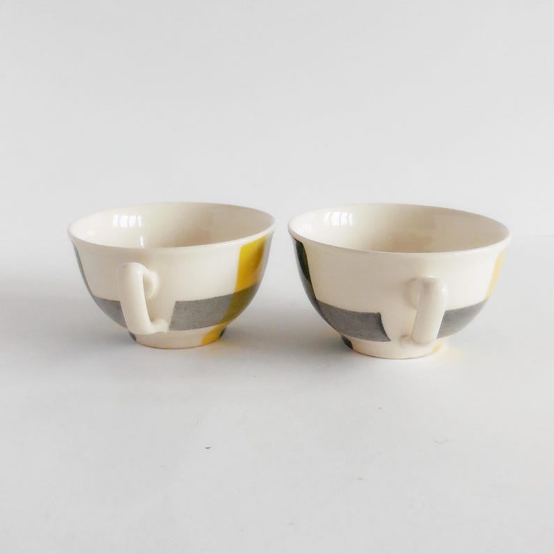 May include: Two white ceramic teacups with handles. Each cup features a design of yellow and gray geometric shapes. The cups are set against a plain white background, highlighting their design and form. Ideal for serving tea or coffee.