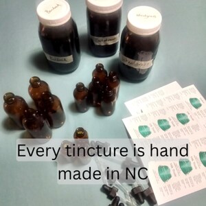 Peut inclure: Plusieurs flacons et bocaux en verre ambr&eacute; remplis d'un liquide fonc&eacute;. L'image comprend des bocaux &eacute;tiquet&eacute;s "Burdock", "Dutystream" et "Wheatgrass". Le texte "Every tincture is hand made in NC" est &eacute;galement visible.