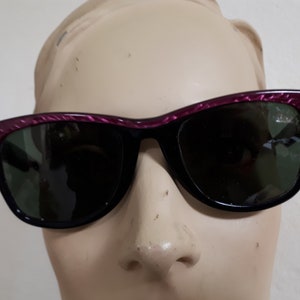 May include: Black sunglasses with a pink and black patterned frame. The sunglasses have green lenses.