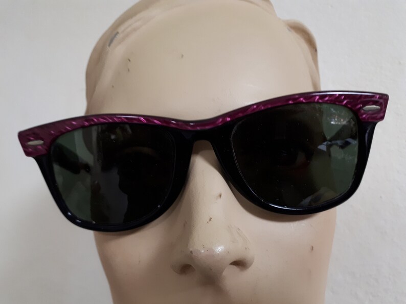 May include: Black sunglasses with a pink and black patterned frame. The sunglasses have green lenses.