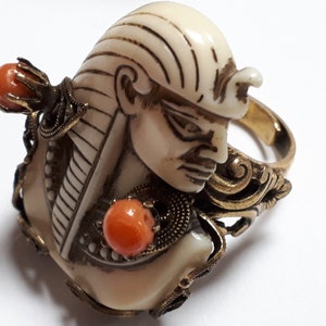 Peut inclure: Une bague en or avec un buste en ivoire sculpté d'un pharaon portant un heaume. La bague a deux perles de corail de chaque côté du buste.
