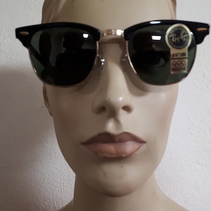 May include: Black and gold-rimmed sunglasses with green lenses. The sunglasses have a classic design with a black upper frame and gold-colored metal accents. A small sticker with text is visible on the lens.