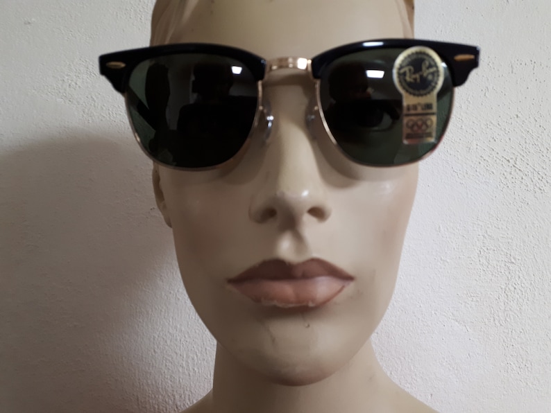 May include: Black and gold-rimmed sunglasses with green lenses. The sunglasses have a classic design with a black upper frame and gold-colored metal accents. A small sticker with text is visible on the lens.