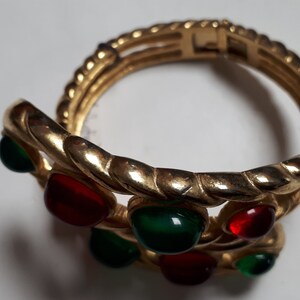 Puede incluir: Un brazalete dorado con un dise&ntilde;o trenzado y piedras preciosas rojas y verdes.