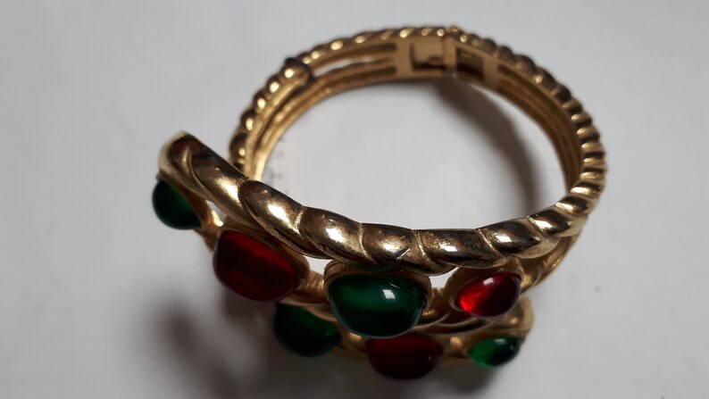 Puede incluir: Un brazalete dorado con un dise&ntilde;o trenzado y piedras preciosas rojas y verdes.