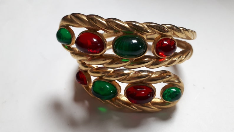 Puede incluir: Un brazalete de oro con un dise&ntilde;o de cuerda trenzada y piedras de vidrio rojo y verde.