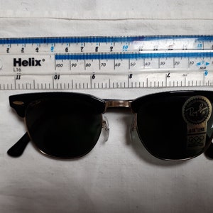 May include: Black Ray-Ban sunglasses with a gold-colored bridge and arms. The sunglasses have a classic design with a black upper frame and dark lenses. A ruler is placed above the sunglasses, measuring in inches and centimeters.