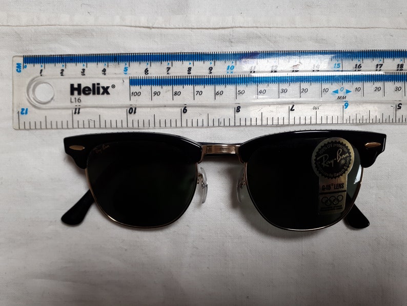 May include: Black Ray-Ban sunglasses with a gold-colored bridge and arms. The sunglasses have a classic design with a black upper frame and dark lenses. A ruler is placed above the sunglasses, measuring in inches and centimeters.