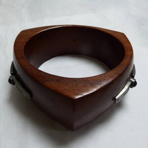 May include: A dark brown wooden bracelet with a square design. The bracelet has a smooth, polished finish and is accented with silver-colored metal details on each side. The bracelet is a bold, statement piece.