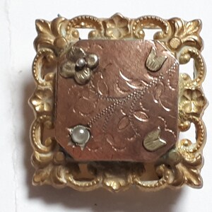May include: A gold-toned brooch with a rectangular, engraved, copper-colored center. The center has a floral design and three small, white pearl-like accents. The brooch is surrounded by a decorative, ornate, gold-toned frame.