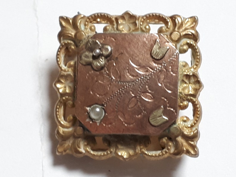 May include: A gold-toned brooch with a rectangular, engraved, copper-colored center. The center has a floral design and three small, white pearl-like accents. The brooch is surrounded by a decorative, ornate, gold-toned frame.