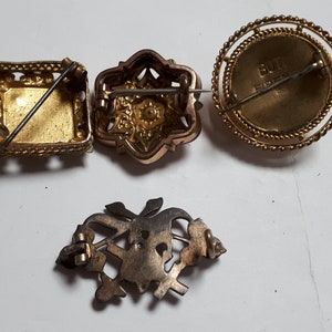May include: Four vintage brooches with intricate designs. One is square with a gold finish, another is a floral design with a rose gold finish, another is round with a gold finish and the text "BUTT FIRE" on the back, and the last is a silver floral design.
