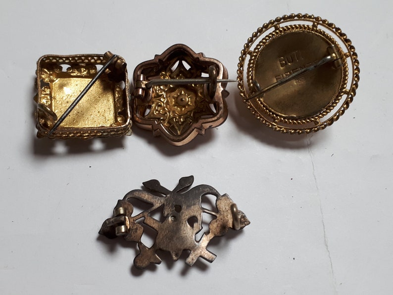 May include: Four vintage brooches with intricate designs. One is square with a gold finish, another is a floral design with a rose gold finish, another is round with a gold finish and the text "BUTT FIRE" on the back, and the last is a silver floral design.