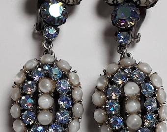 May include: A pair of vintage clip-on earrings. Each earring features a cluster of three white, round stones at the top, followed by a large, iridescent crystal. The oval-shaped drop is adorned with white and blue-toned stones.