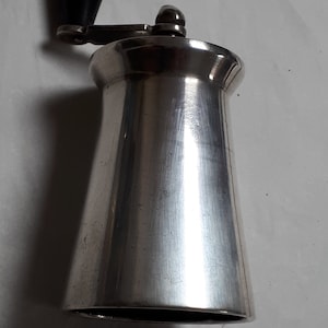 Può includere: Un macinino da pepe argentato con una manopola in plastica nera sulla parte superiore. Il macinino è in metallo e ha una finitura liscia e lucida.