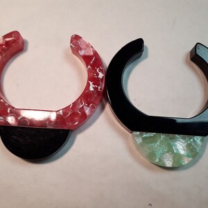 Può includere: Due braccialetti in resina, uno con un designo marmorizzato rosso e bianco e un fondo nero, l'altro con una parte superiore nera e un fondo marmorizzato verde e bianco.
