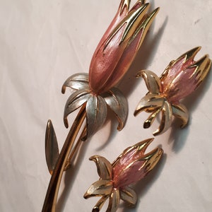 以下が含まれることがあります： ピンクと白の花のデザインが施された、ゴールドトーンのブローチとイヤリングのセット。花はゴールドトーンの茎と葉を持っています。