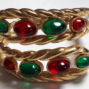 Puede incluir: Un brazalete de oro con un dise&ntilde;o de cuerda trenzada y piedras cabuj&oacute;n rojas y verdes.