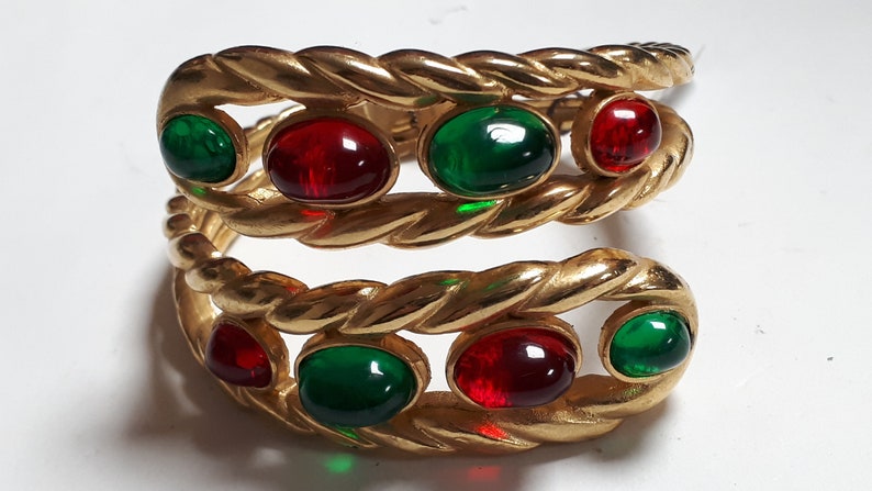 Puede incluir: Un brazalete de oro con un dise&ntilde;o de cuerda trenzada y piedras cabuj&oacute;n rojas y verdes.
