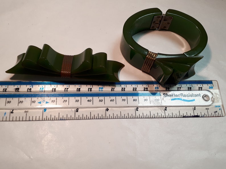 May include: A set of two green Bakelite jewelry pieces. A large green bangle bracelet with a gold metal clasp and a smaller green bow brooch with a gold metal accent.