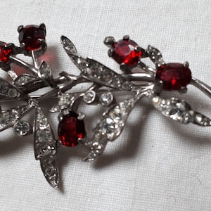 Op de afbeelding: Een zilverkleurige broche met een bladontwerp en rode strasssteentjes.