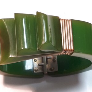 May include: A green Bakelite bangle bracelet with a gold-toned metal clasp and a decorative, geometric design on the front.