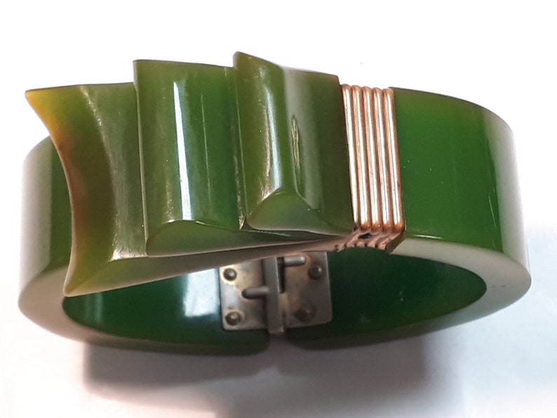 May include: A green Bakelite bangle bracelet with a gold-toned metal clasp and a decorative, geometric design on the front.