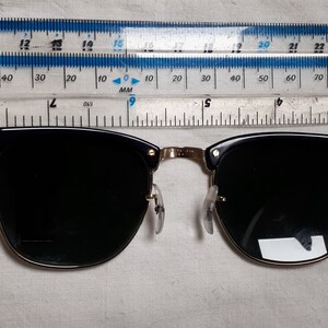 May include: Black and gold-rimmed sunglasses with dark lenses. The sunglasses have a classic design with a black upper frame and gold-colored accents. The lenses are dark, providing shade. The glasses are measured with a ruler.