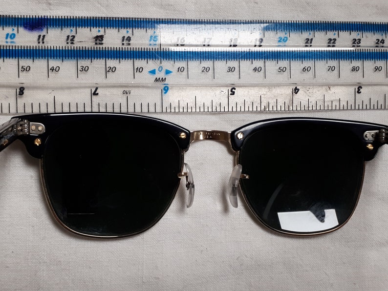 May include: Black and gold-rimmed sunglasses with dark lenses. The sunglasses have a classic design with a black upper frame and gold-colored accents. The lenses are dark, providing shade. The glasses are measured with a ruler.