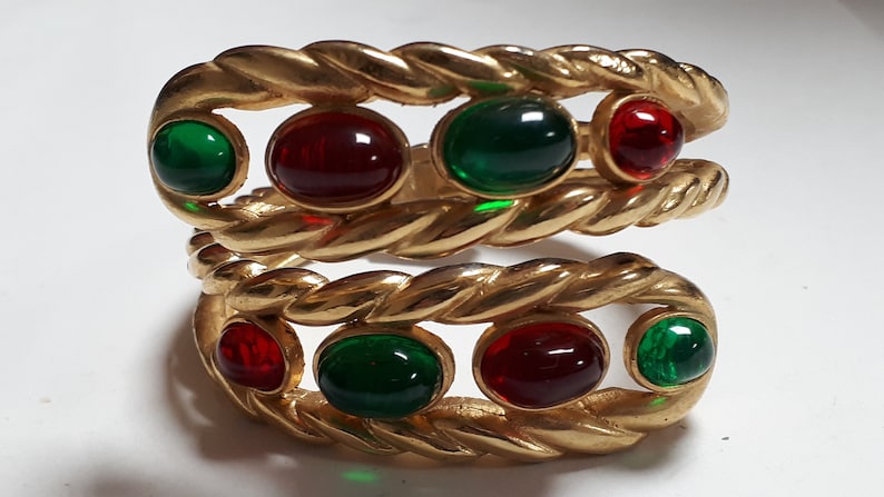 Puede incluir: Un brazalete de oro con un dise&ntilde;o de cuerda trenzada y piedras cabuj&oacute;n rojas y verdes alternas.