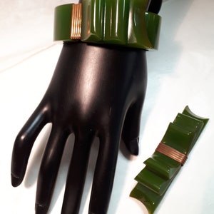 May include: A green and gold plastic bracelet with a bow design. The bracelet is on a black mannequin hand. There is a matching green and gold plastic brooch next to the hand.