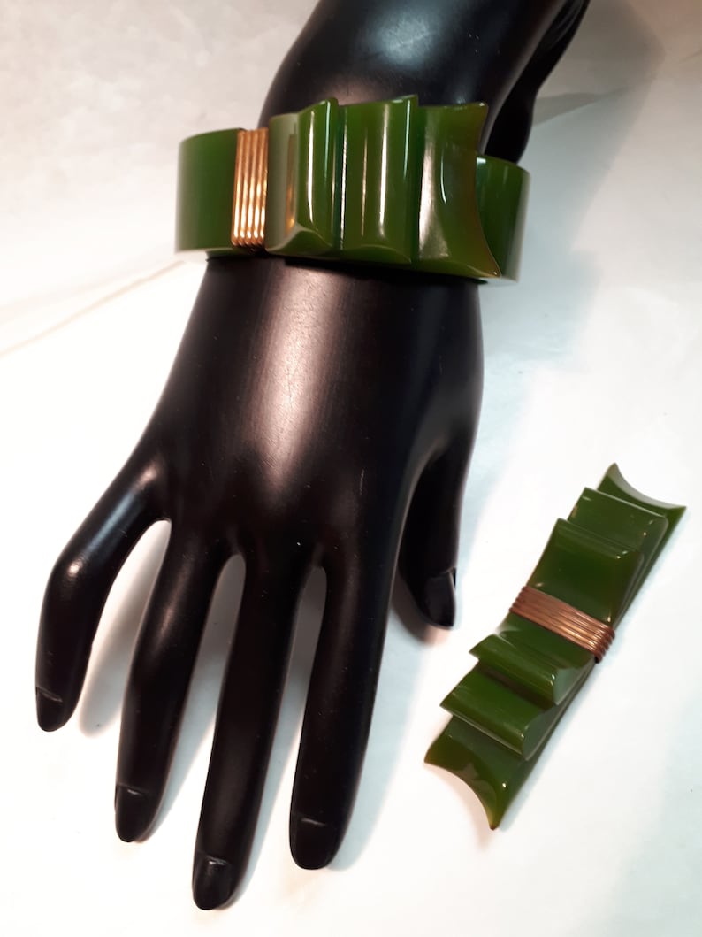 May include: A green and gold plastic bracelet with a bow design. The bracelet is on a black mannequin hand. There is a matching green and gold plastic brooch next to the hand.