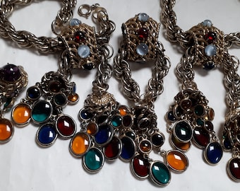 May include: A gold chain necklace with a cluster of colorful glass stones hanging from the bottom. The necklace has two matching earrings with purple stones.
